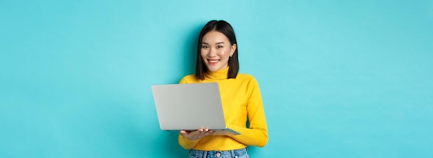 Mujer asiática feliz en jersey amarillo usando una computadora portátil comprando en línea o trabajando de pie sobre la espalda azul