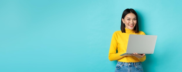 Mujer asiática feliz en jersey amarillo usando una computadora portátil comprando en línea o trabajando de pie sobre la espalda azul