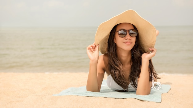 Mujer asiática feliz joven con el sunhat que miente en la playa en verano