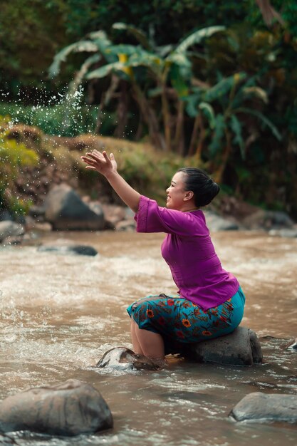 Mujer asiática feliz jugando con el agua cerca del río mientras se sienta en la roca y lleva un vestido morado