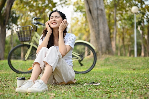 Mujer asiática feliz con los ojos cerrados sentada en el césped escuchando música relajándose en el parque