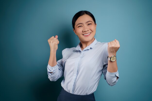 Mujer asiática feliz de pie seguro mostrando su puño hacer un gesto ganador aislado en azul.