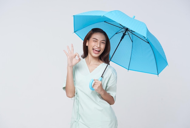 Mujer asiática feliz de pie sosteniendo un paraguas azul y mostrando el signo de ok aislado en el fondo blanco