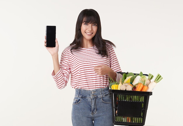 Mujer asiática feliz que muestra un teléfono inteligente con una cesta llena de comestibles de verduras frescas