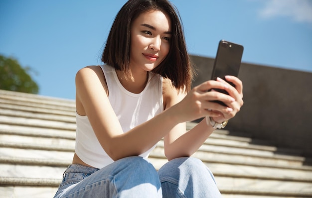 Mujer asiática feliz sentada en las escaleras de la calle y tomando selfie sonriendo a la cámara del teléfono móvil Chica chateando en línea mientras descansa al aire libre