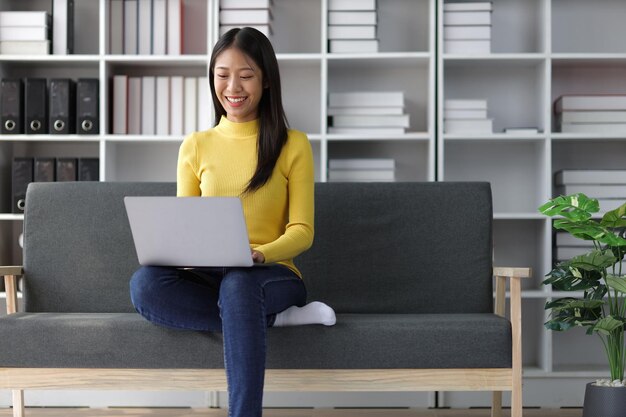 Mujer asiática feliz sentada en el sofá y usando una computadora portátil en la sala de estar en casa