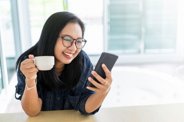 Mujer asiática feliz sonriendo con smartphone y taza de café