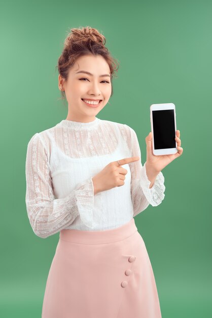 Mujer asiática feliz sorprendida que muestra la pantalla del smartphone en blanco y que señala en él. Fondo verde aislado.