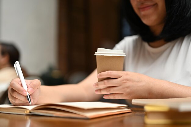 Una mujer asiática feliz tomando café y escribiendo un diario mientras se relaja en una cafetería