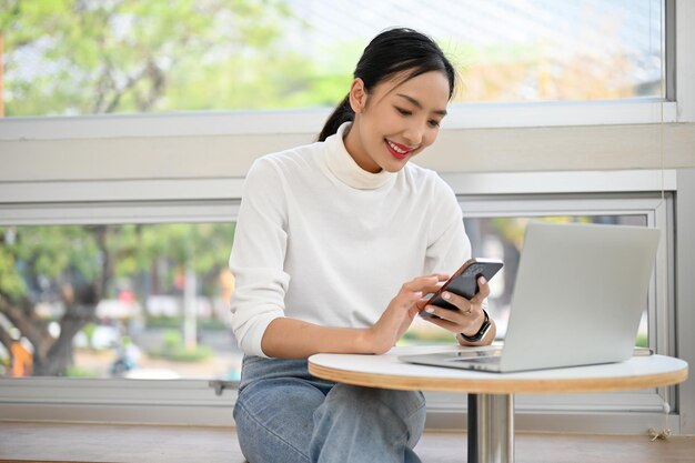 Mujer asiática feliz trabajando remotamente en el café usando su teléfono inteligente sentado en la mesa