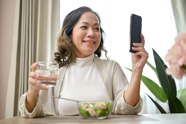 Mujer asiática feliz usando su teléfono inteligente mientras desayuna ensalada saludable