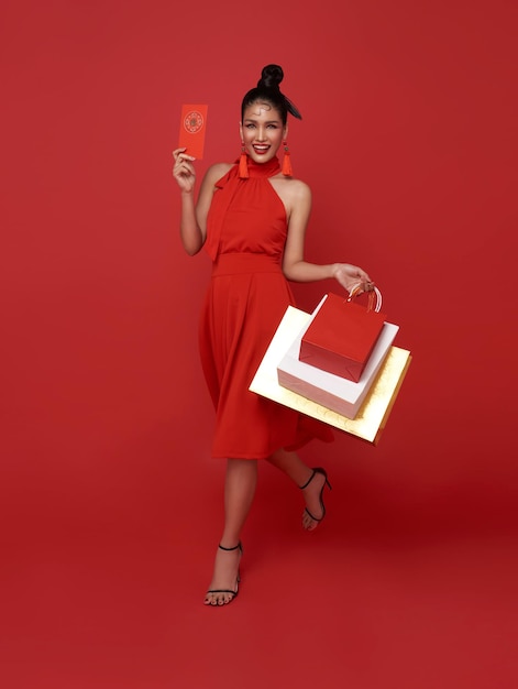 Mujer asiática feliz con vestido rojo sosteniendo bolsas de compras y sobres rojos con cupones de regalo