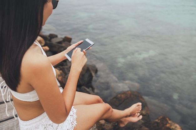 Mujer asiática feliz viajero con teléfono móvil y relajarse en el puente de madera en la playa
