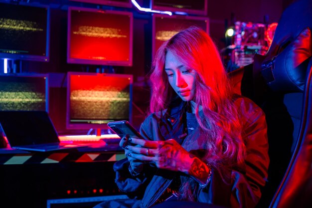 Mujer asiática frente a una pantalla de monitor de computadora usando un teléfono móvil con luz de neón hacker usar negro