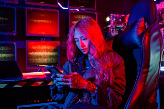 Mujer asiática frente a una pantalla de monitor de computadora usando un teléfono móvil con luz de neón hacker usar negro