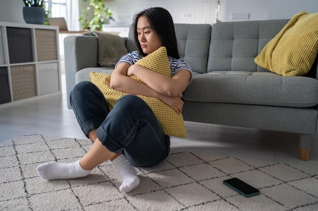 Una mujer asiática frustrada sentada en el suelo cerca del sofá abrazando una almohada piensa en problemas de relación