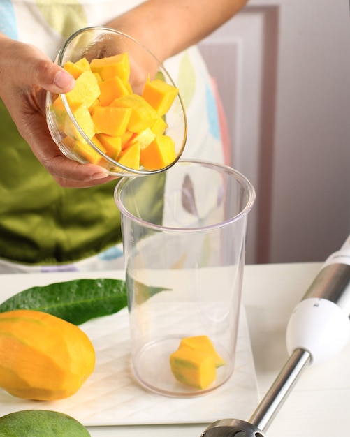 Foto mujer asiática haciendo jugo de mango con batidora de mano. paso a paso, coloque los dados de mango en el tazón de la licuadora