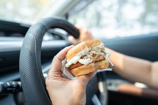 Una mujer asiática con una hamburguesa para comer en el coche es peligrosa y arriesga un accidente.