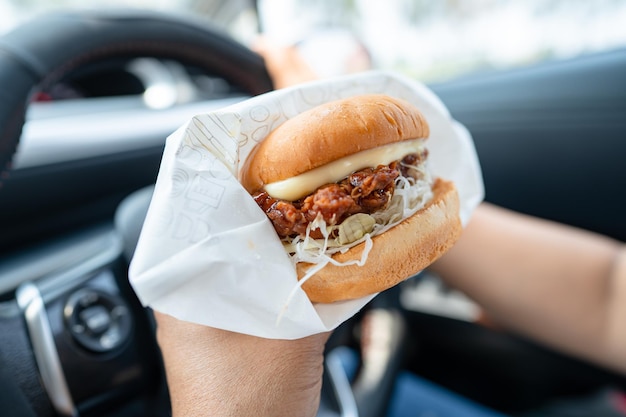 Una mujer asiática con una hamburguesa para comer en el coche es peligrosa y arriesga un accidente.