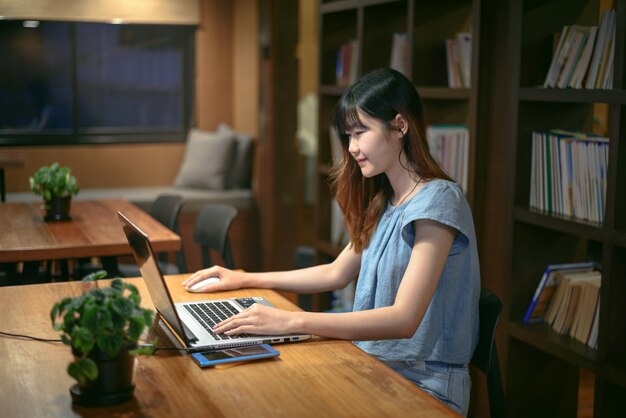 Mujer asiática hermosa feliz del estudiante que trabaja en el ordenador portátil en sitio moderno de la biblioteca