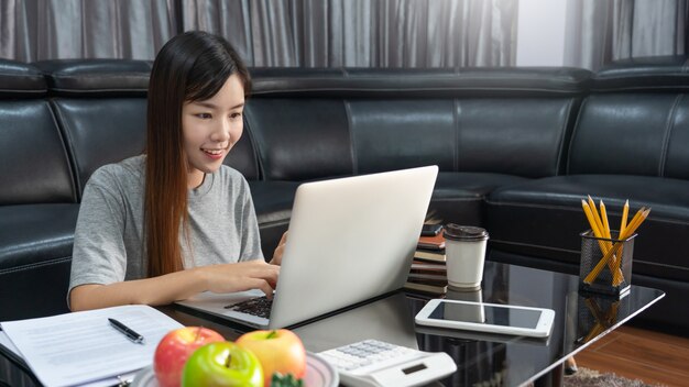 Mujer asiática hermosa joven atractiva Empresario o profesional independiente que trabaja en casa con los informes comerciales del ordenador portátil y las comunicaciones en línea en el sofá de la sala de estar, trabajando concepto de acceso remoto.