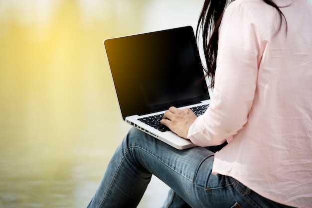 Mujer asiática hermosa joven con la cara sonriente que trabaja al aire libre en un parque público. Trabajando en la computadora portátil al aire libre. Imagen recortada de la mujer que trabaja en la computadora portátil mientras está sentado en un parque.