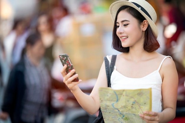 Mujer asiática hermosa joven que mira la pantalla del smartphone y que sostiene el mapa en su mano