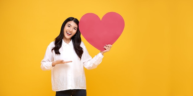 Mujer asiática hermosa joven que muestra la muestra del corazón rojo en amarillo. amor y feliz concepto de San Valentín.