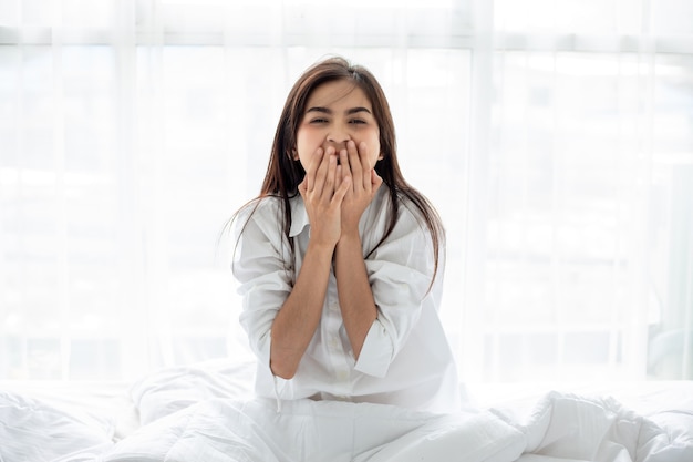 Mujer asiática Hermosa joven sonriente sentada y durmiendo en la cama blanca y estirándose por la mañana en el dormitorio después de despertarse en su cama completamente descansada