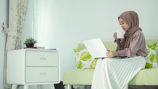 Mujer asiática en hijab trabajando en casa sentado relajado disfrutando de una bebida