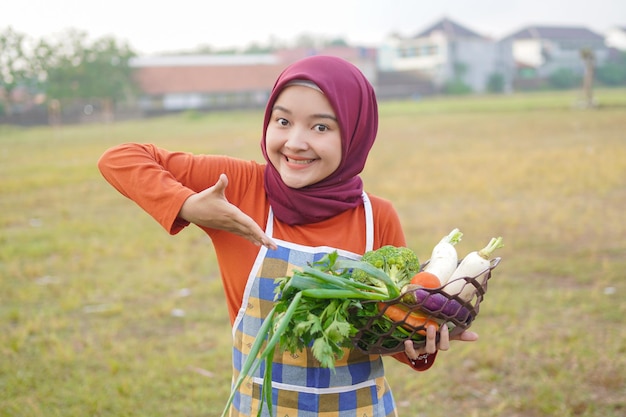 Foto mujer asiática hijab usa delantal, sostiene verduras y señala algo