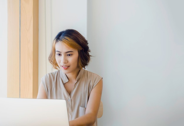 Mujer asiática independiente con cabello corto de moda mirando la pantalla mientras trabaja con una computadora portátil en un fondo de pared blanca con espacio para copiar cerca del vidrio de la ventana en el café