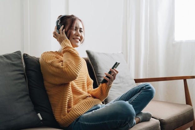 Mujer asiática independiente con teléfono móvil escuchando música en auriculares y relájese en casa Niña feliz sentada en el sofá en la sala de estar