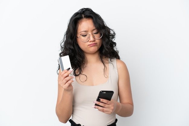 Mujer asiática joven aislada sobre fondo blanco comprando con el móvil con una tarjeta de crédito mientras piensa
