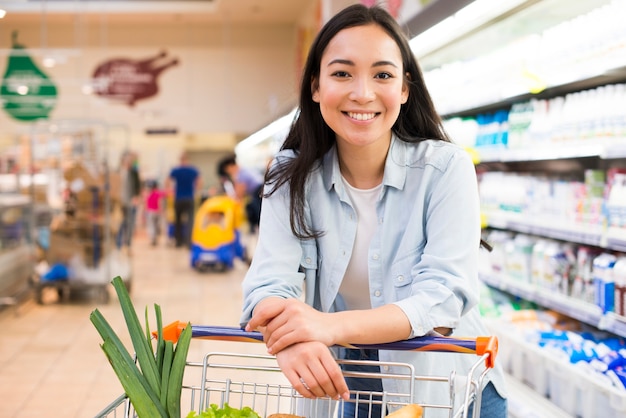 Mujer asiática joven alegre con el carro de compras en el supermercado