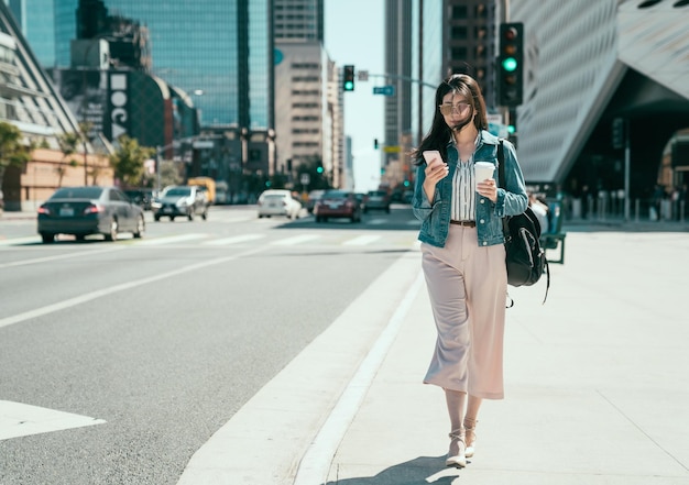 mujer asiática joven alegre de longitud completa que usa teléfono móvil en la calle soleada de la ciudad y bebe café para llevar en una taza de papel. Empleada con mochila caminando por la carretera en el ajetreado tráfico de la zona de negocios