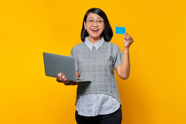 Mujer asiática joven alegre que sostiene la computadora portátil y la tarjeta de crédito en fondo amarillo