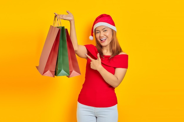 Mujer asiática joven alegre en sombrero de Papá Noel que señala el dedo en la bolsa de compras sobre fondo amarillo