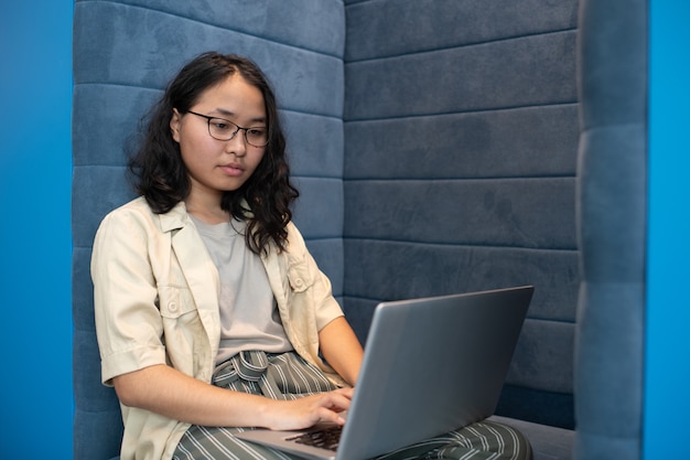 Mujer asiática joven en anteojos y ropa casual usando laptop