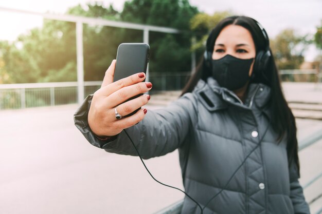 Mujer asiática joven con auriculares y mascarilla tomando selfie con teléfono móvil al aire libre. Concepto de estilo de vida. Concepto de tecnología.