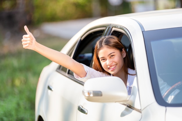 Mujer asiática joven en la camisa blanca que conduce su coche y que muestra los pulgares para arriba