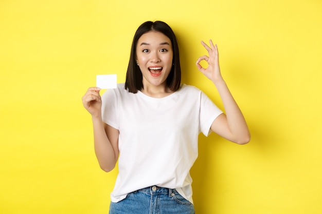 Mujer asiática joven en camiseta blanca casual que muestra una tarjeta de crédito plástica y un gesto bien, recomendar banco, sonriendo a la cámara, fondo amarillo