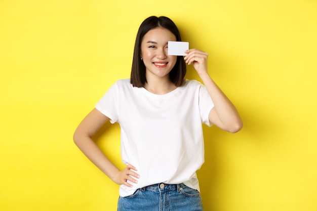 Mujer asiática joven en camiseta blanca casual que muestra la tarjeta de crédito plástica y sonriendo a la cámara, fondo amarillo