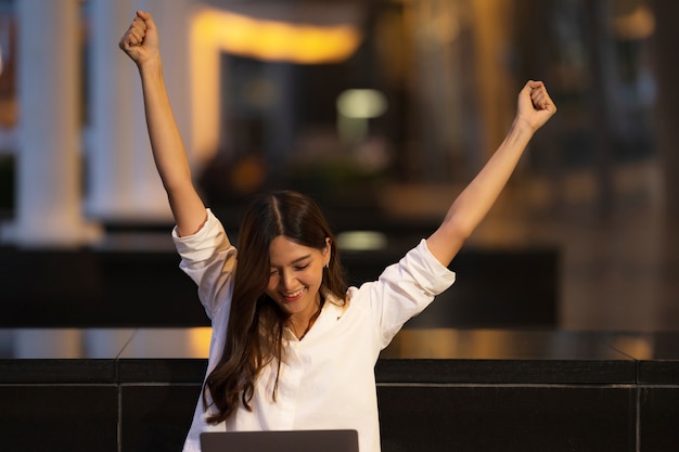 Mujer asiática joven con cara de sorpresa gritando feliz usando la computadora portátil en una ciudad por la noche