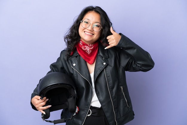 Mujer asiática joven con un casco de motocicleta aislado sobre fondo púrpura haciendo gesto de teléfono. Llámame señal