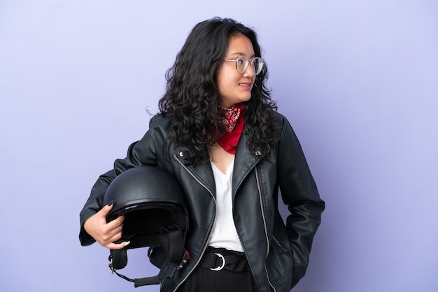 Mujer asiática joven con un casco de motocicleta aislado sobre fondo púrpura mirando de lado