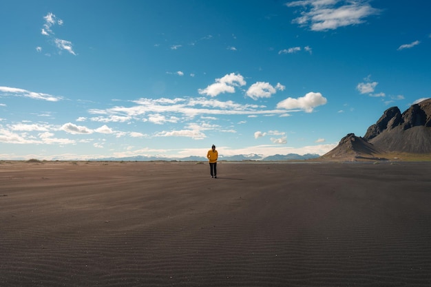 Mujer asiática joven en chaqueta amarilla caminando en la playa de arena negra en un día soleado en Vestrahorn Islandia