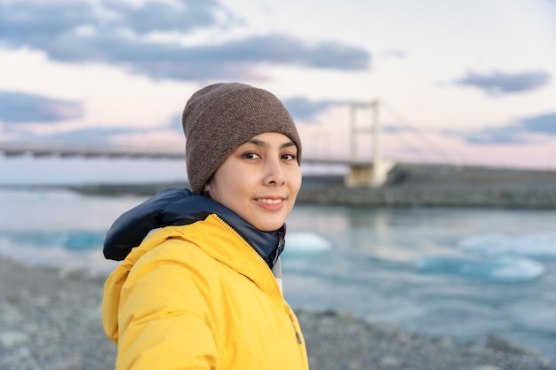 Mujer asiática joven en chaqueta amarilla con gorro de pie y sonriendo en la laguna glaciar en Islandia