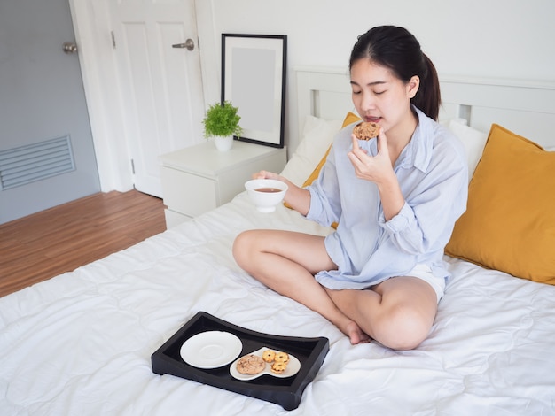 La mujer asiática joven come la galleta del café en cama