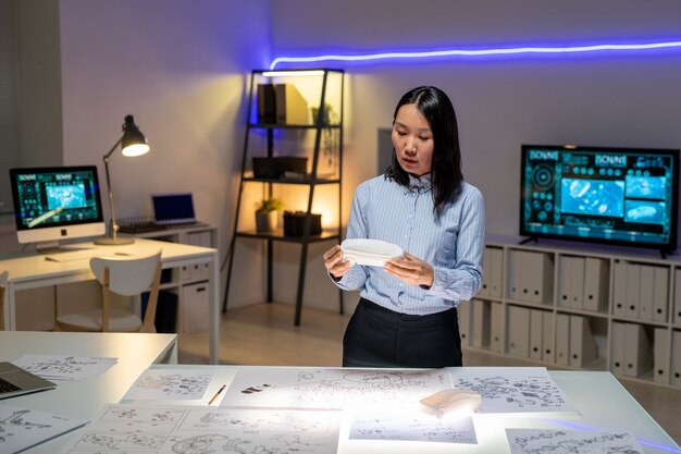 Mujer asiática joven concentrada en ropa formal de pie en el escritorio con diseños y sosteniendo maqueta mientras produce el diseño del coche en la oficina